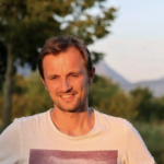 Smiling man with short brown hair wearing a white T-shirt stands outdoors with a blurred background of trees and mountains in soft, warm light.